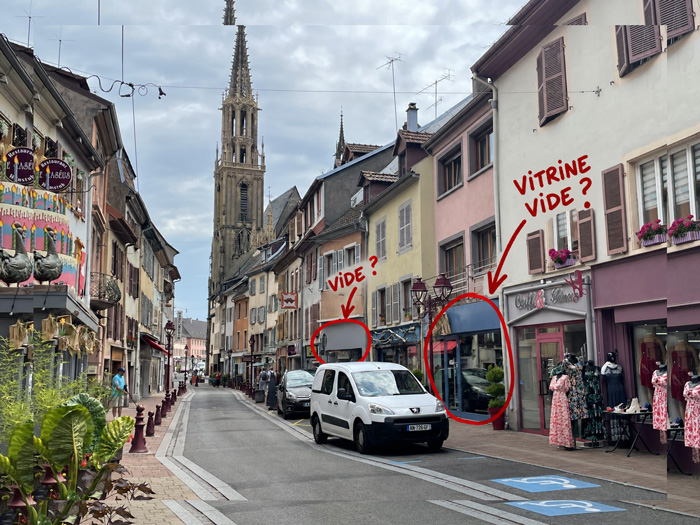 Rue commerçante du centre-ville de Thann avec la collégiale Saint-Thiébaut en arrière-plan et des annotations manuscrites rouges sur des vitrines